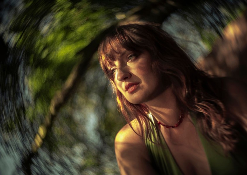 A close up image of a woman with wavy brown hair wearing a red necklace and green topx
